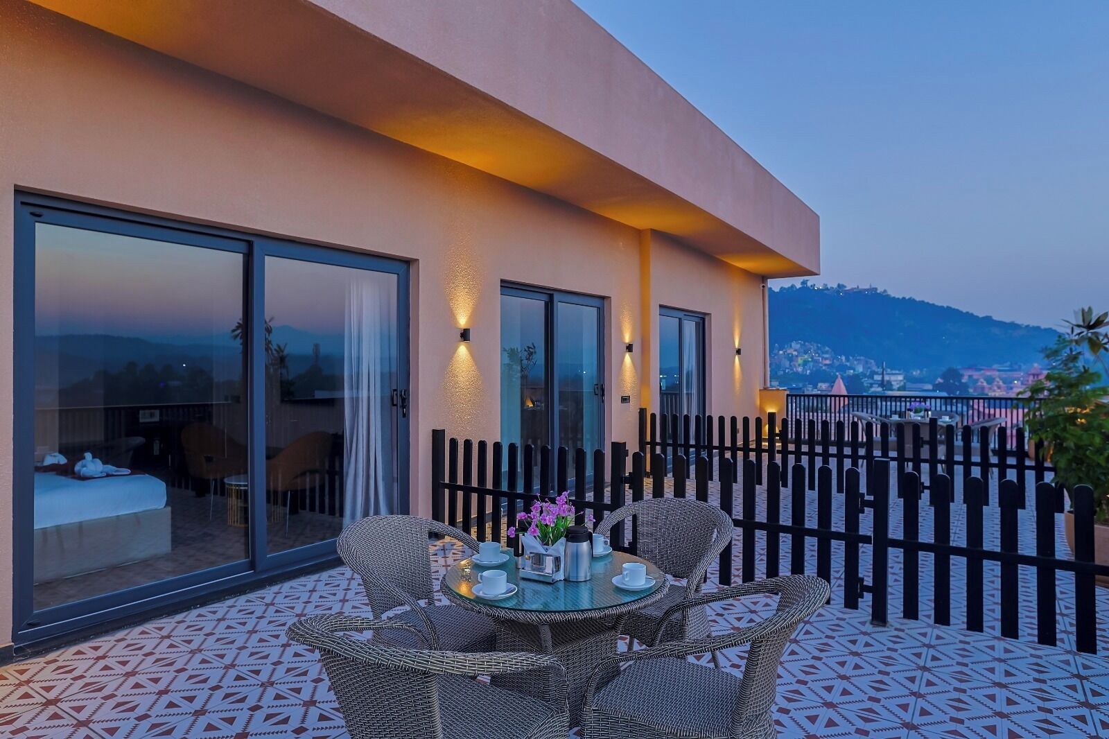 Une chambre d’hôtel moderne avec balcon, meublée de chaises et d’une table en osier pour deux personnes, offrant une vue panoramique.