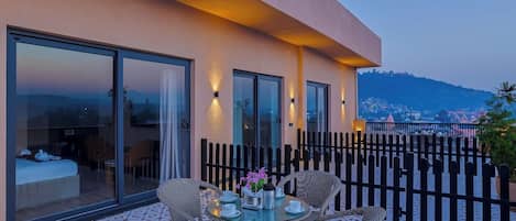 A modern hotel room with a balcony, furnished with wicker chairs and a table set for two, overlooking a scenic view.