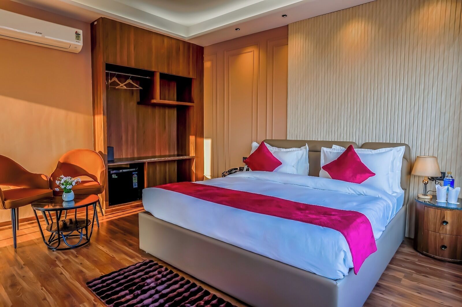A hotel room with a large bed, a wooden wardrobe, a seating area with a chair and table, and a bedside table with a lamp.