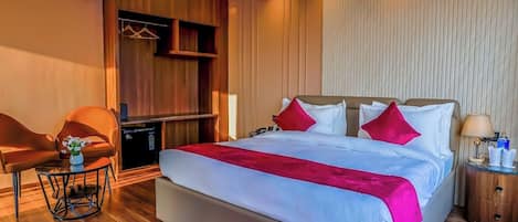 A hotel room with a large bed, a wooden wardrobe, a seating area with a chair and table, and a bedside table with a lamp.