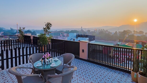 A rooftop terrace with a table and chairs, potted plants, and a view of the cityscape during sunset. - Arina Vedic Haveli 2 Min walk to Ganga (Haridwar)