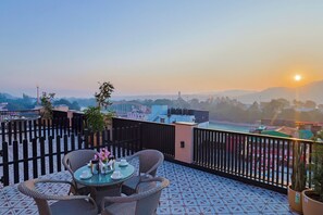 Une terrasse sur le toit, avec une table et des chaises, des plantes en pot et une vue sur le paysage urbain au coucher du soleil.