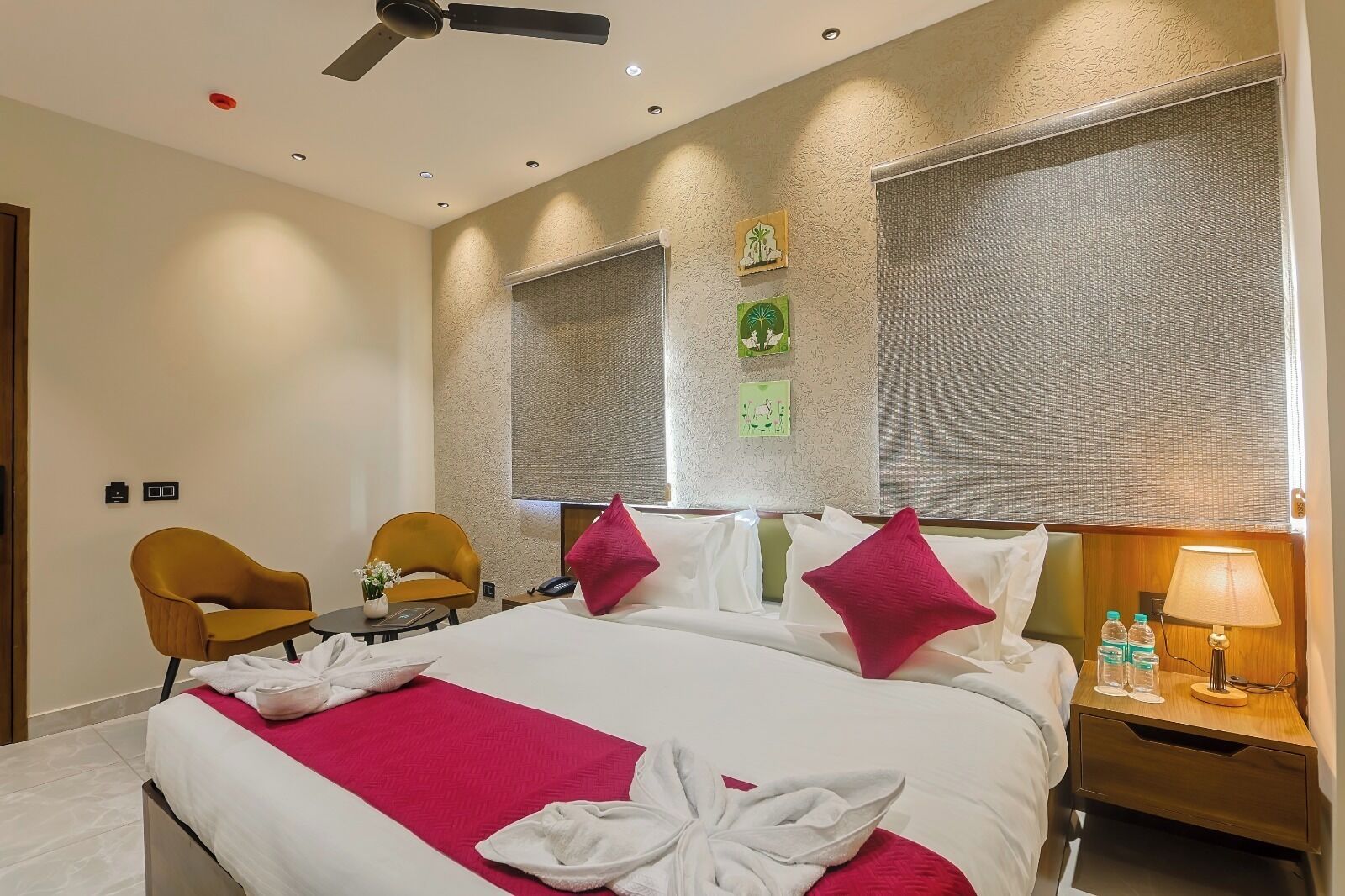 A hotel room with a bed, two chairs, a desk, a nightstand, a ceiling fan, and a textured wall.