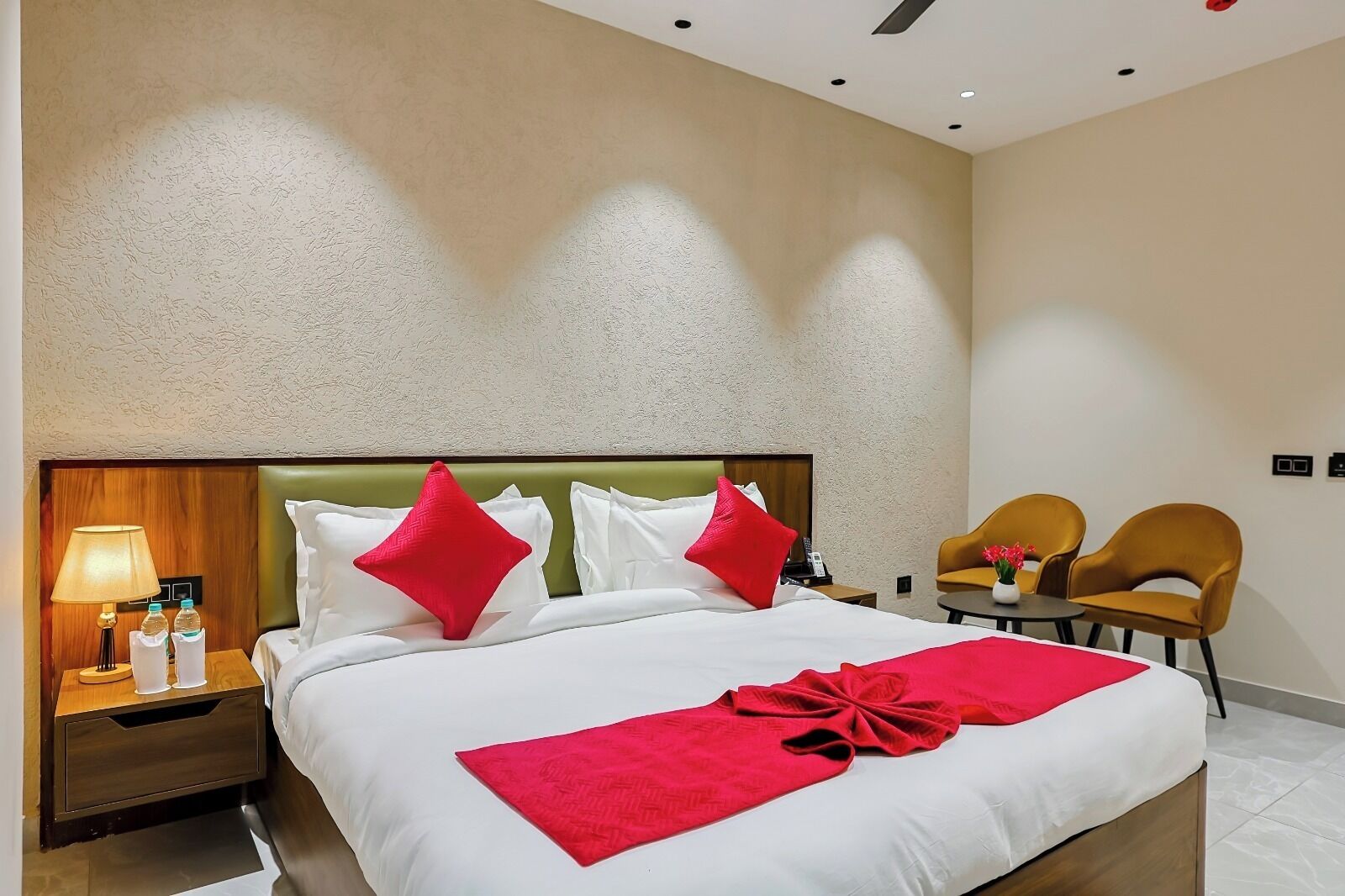 A hotel room with a large bed, two red pillows, a bedside table with a lamp and water bottles, and two armchairs.