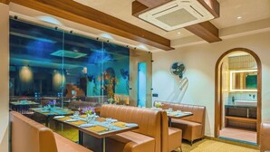 A restaurant with booth seating, a large window with a view of the dining area, and a fan on the wall. - Arina Vedic Haveli 2 Min walk to Ganga (Haridwar)