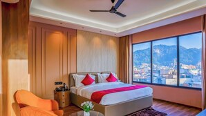 A hotel room with a large bed, a desk, a chair, and a view of a cityscape through a large window. - Arina Vedic Haveli 2 Min walk to Ganga (Haridwar)