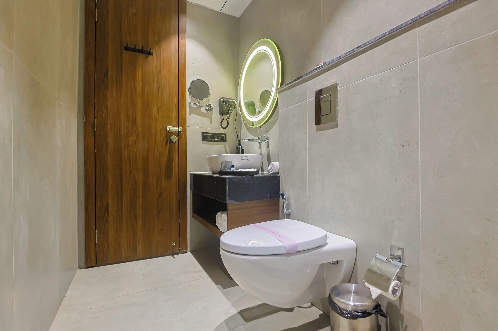 A modern bathroom with a wooden door, a white toilet, a sink with a mirror, and a trash bin.