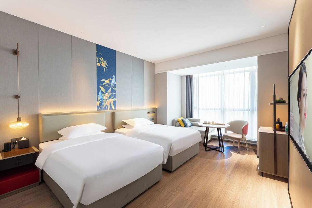 A hotel room with two beds, a desk, a chair, a TV, and a window with curtains.