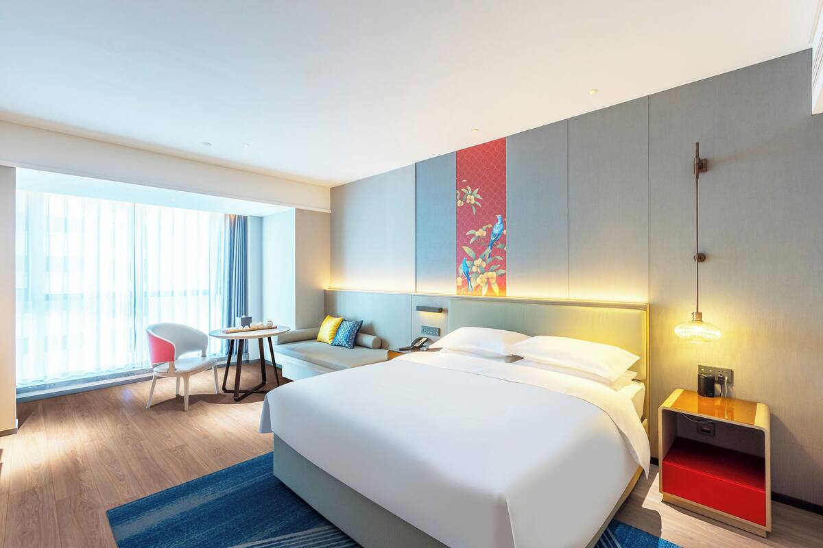 A hotel room with a large bed, a desk, a chair, a small table, and a nightstand.
