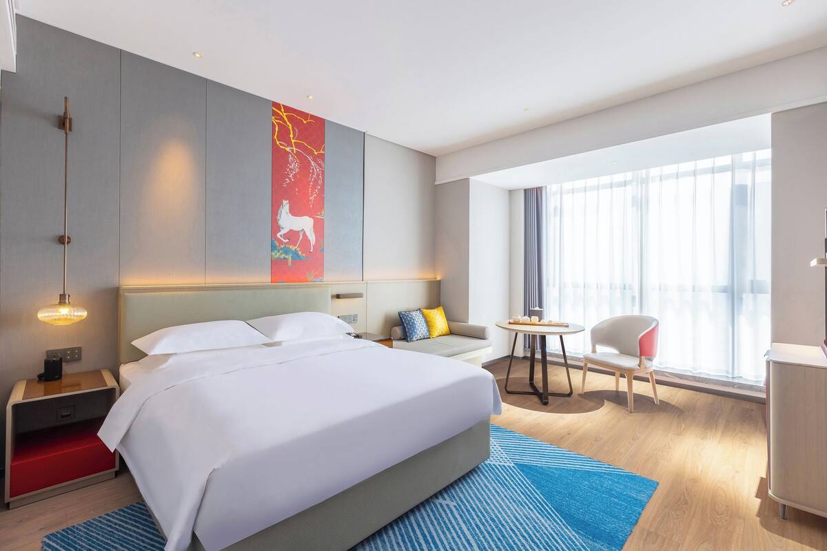 A hotel room with a large bed, a desk, a chair, a small table, and a window with curtains.