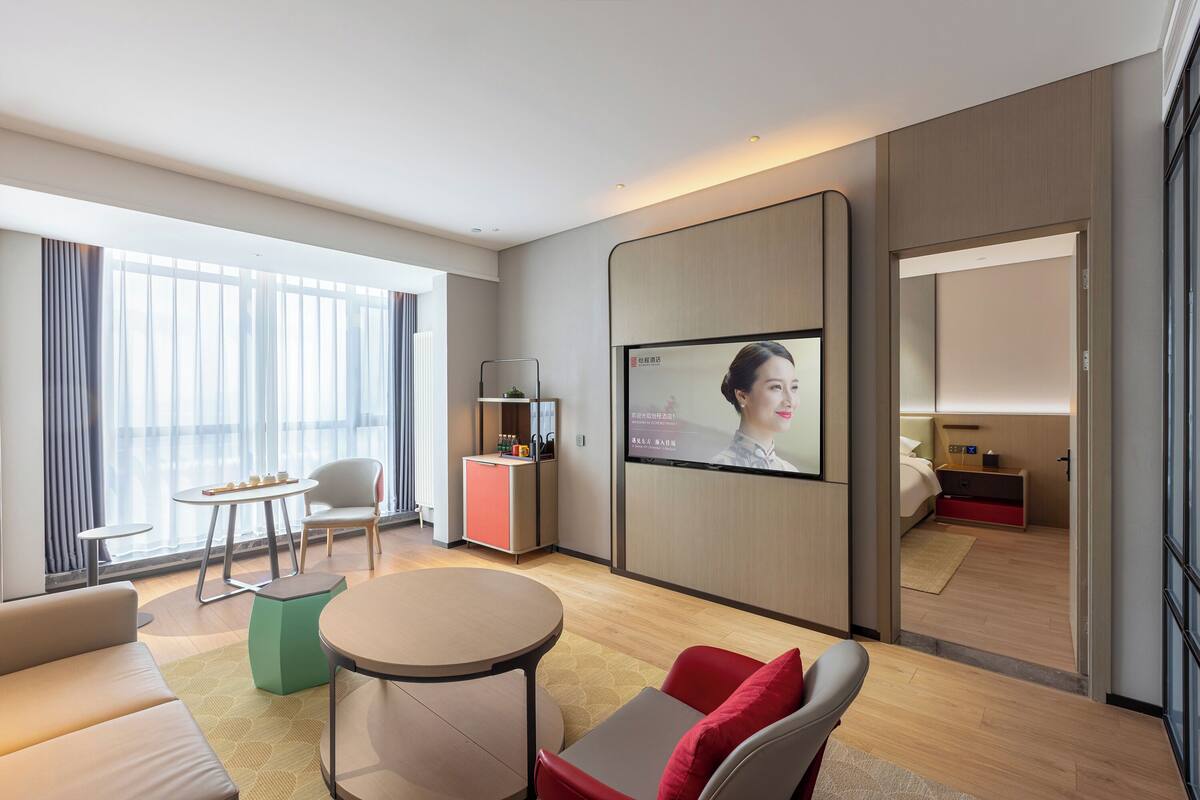 A modern hotel room with a large window, a flat-screen TV, a sofa, a small table, and a cabinet.