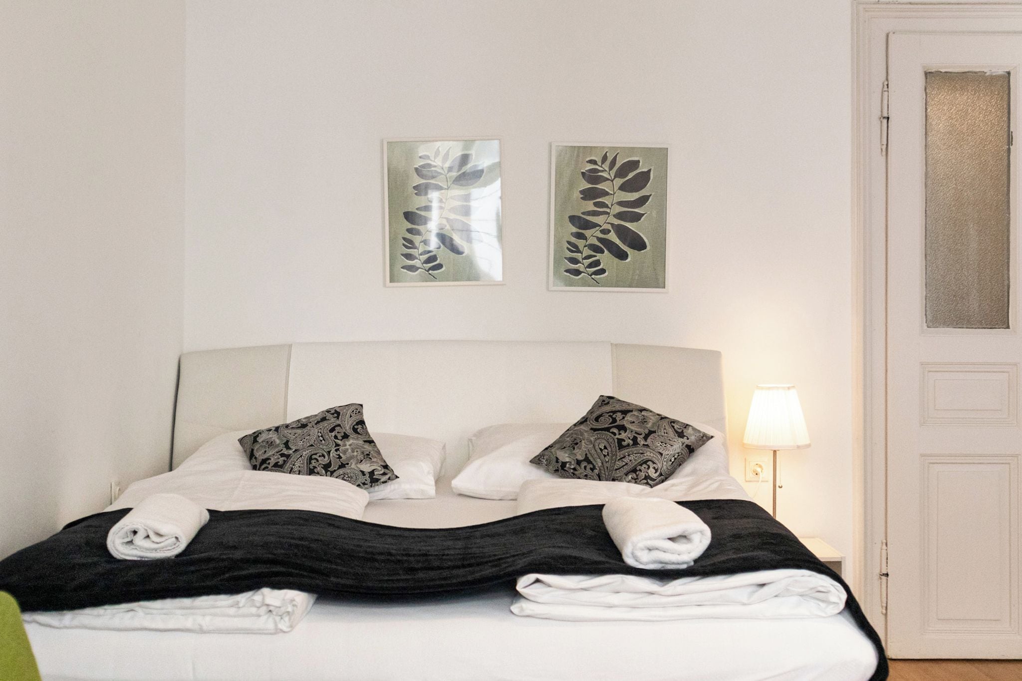 Un lit bien fait, avec du linge de lit blanc et noir, deux coussins à motifs et une serviette pliée posée sur le côté.