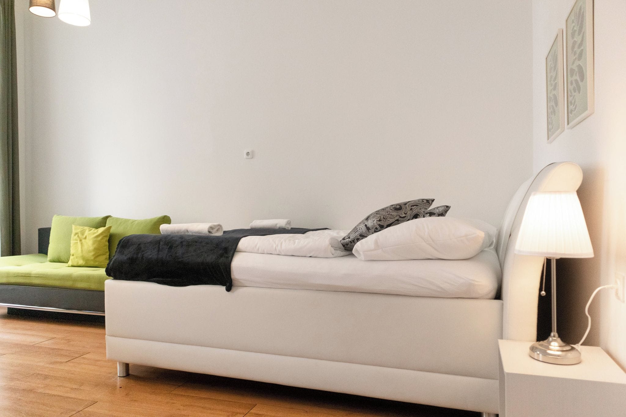 A modern hotel room with a white sofa bed, a green armchair, and a bedside table with a lamp.