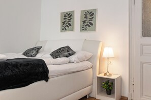 A bed with white bedding and black throw, a bedside lamp, and a small plant on a nightstand. - Charming 2BR Apt Prime City Spot (Vienna)