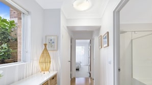 A hallway with white walls, a wooden floor, and a door at the end. There is a framed picture on the wall and a plant on the left. - Sunshine and Sea Breeze (SUNSHINE AND SEA BREEZE)