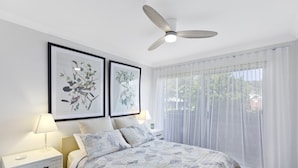 A bedroom with a bed, two framed botanical prints on the wall, a ceiling fan, and a window with sheer curtains. - Sunshine and Sea Breeze (SUNSHINE AND SEA BREEZE)