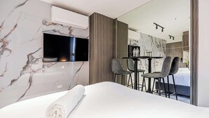 Ein modernes Hotelzimmer mit Marmorwand, einem Flachbildfernseher, einem Bett mit gerolltem weißen Handtuch, einem kleinen Tisch mit zwei Stühlen und Blick in ein Schlafzimmer mit Bett und Nachttisch.