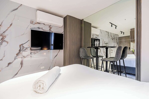 Une chambre d’hôtel moderne avec un mur en marbre, un téléviseur à écran plat, un lit avec une serviette blanche roulée, une petite table avec deux chaises et une vue sur une chambre à coucher avec un lit et une table de chevet.