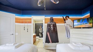 A room with a Star Wars theme, including a Darth Vader figure, a lightsaber, and a TV displaying a beach scene. There are two beds with white bedding and a bathroom visible in the background. - Disney Family Escape - Stunning 5-bed Villa With Private Pool (Kissimmee)