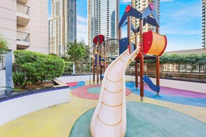 A playground with a slide and climbing structures, surrounded by a pool area and greenery. - FIRST CLASS |1BR| Downtown Elegance | Burj Khalifa (Downtown)
