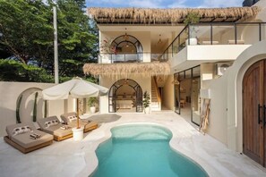 Une villa moderne dotée d’une piscine, de chaises longues et d’un balcon avec toit de chaume.