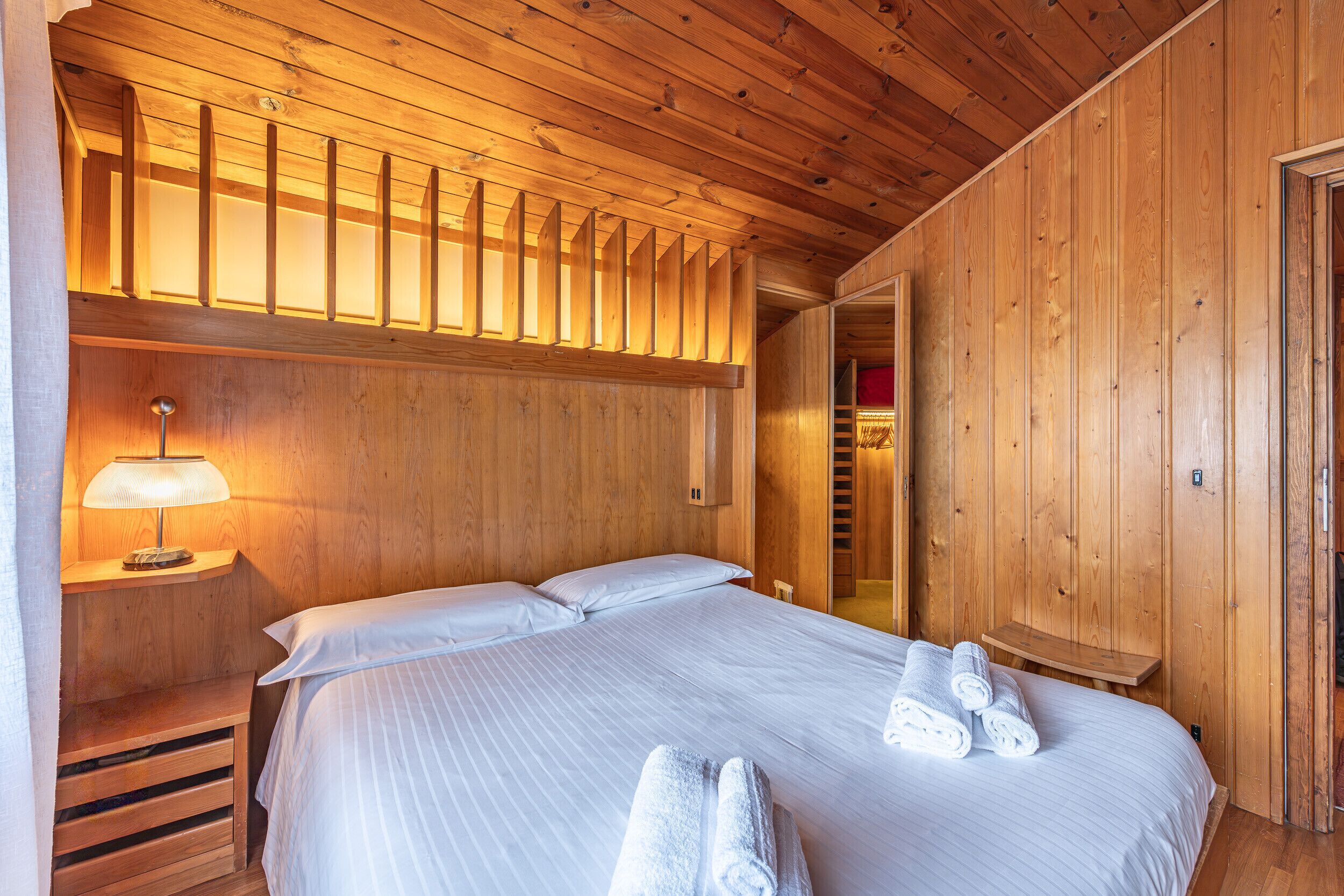 A wooden-paneled room with a bed, a nightstand with a lamp, and a wooden bench.