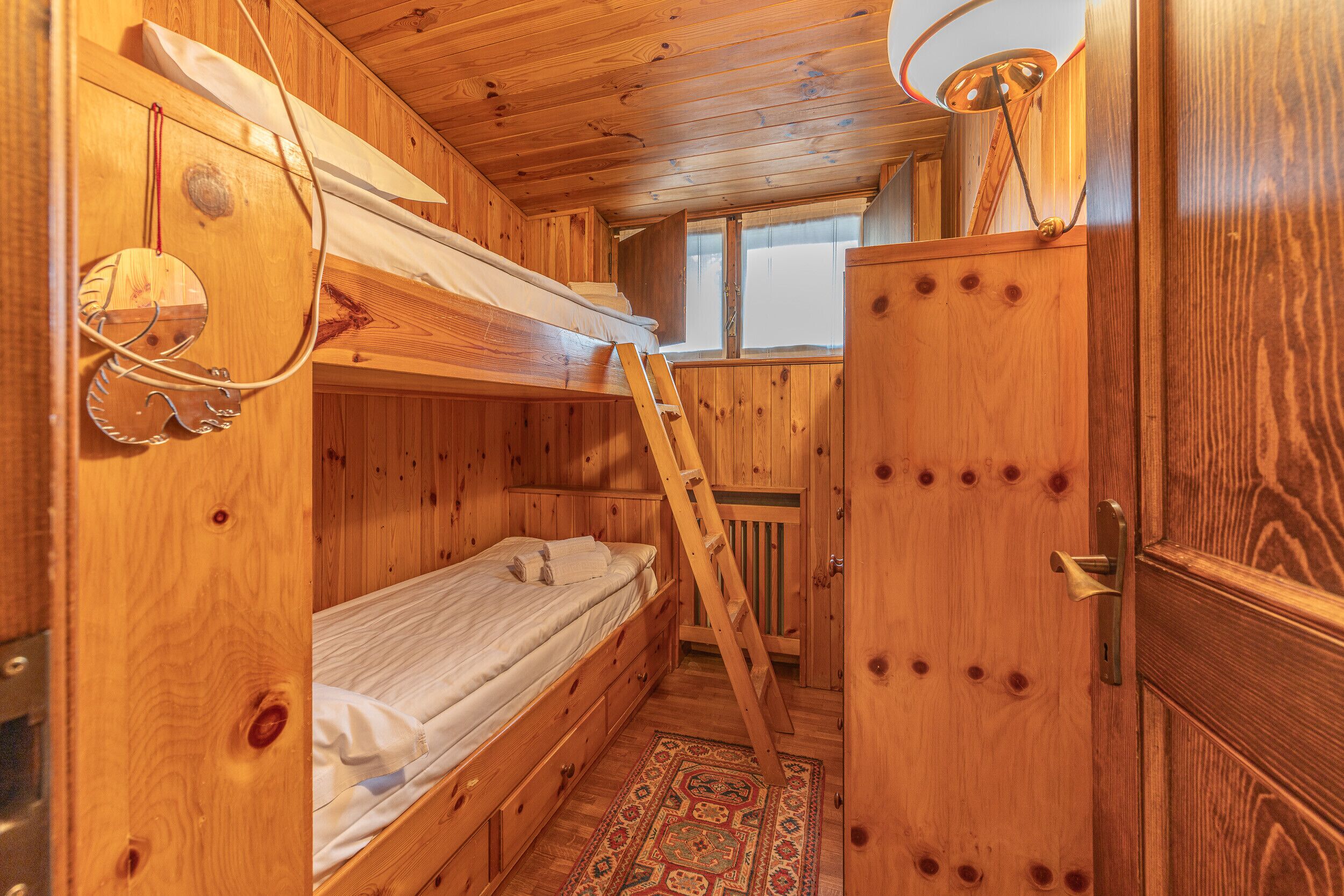 A wooden cabin interior with bunk beds, a ladder, a door with a handle, and a window with a view.