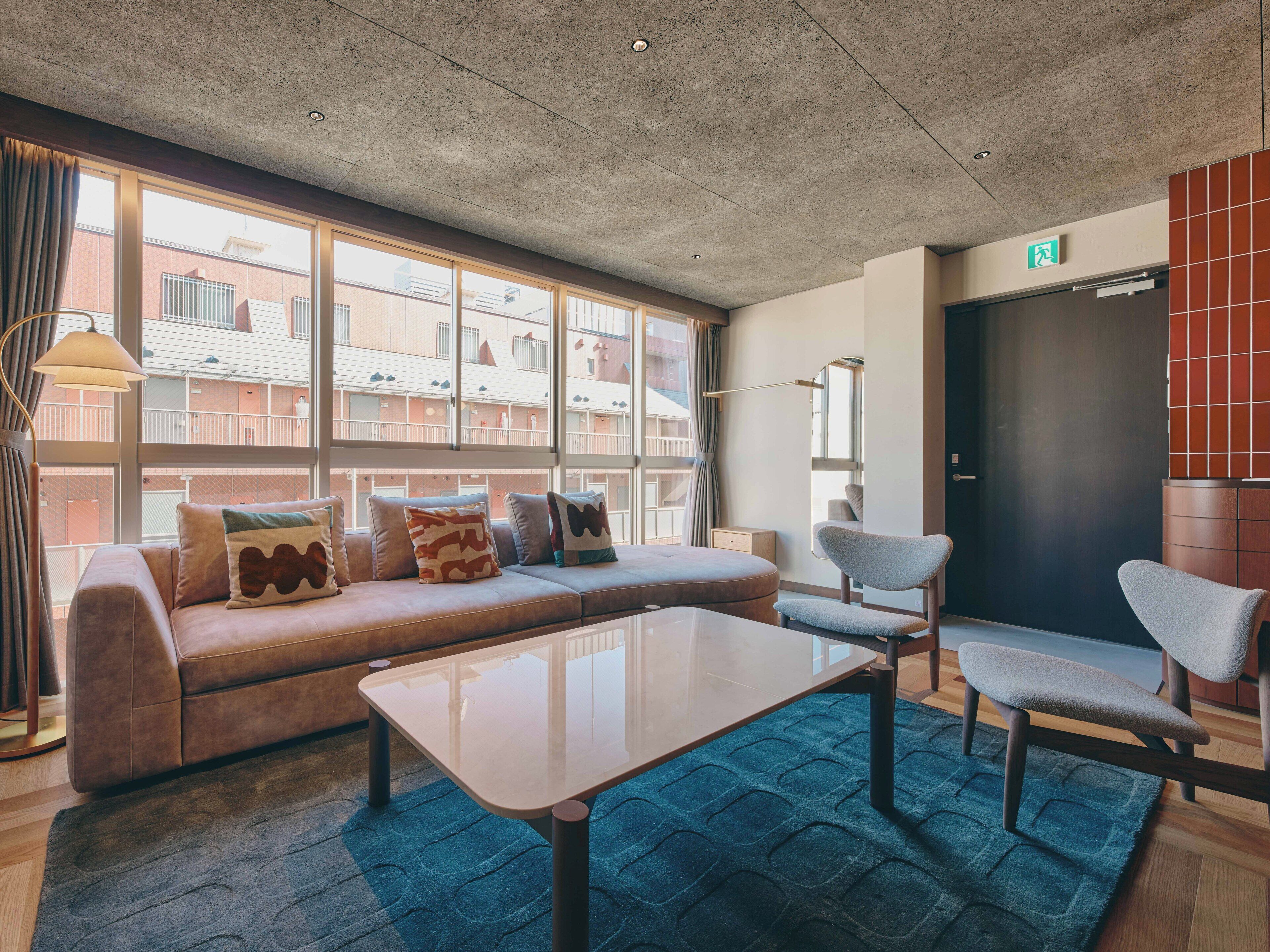 A modern lounge area with a sofa, chairs, a table, and large windows offering a view of the building's exterior.