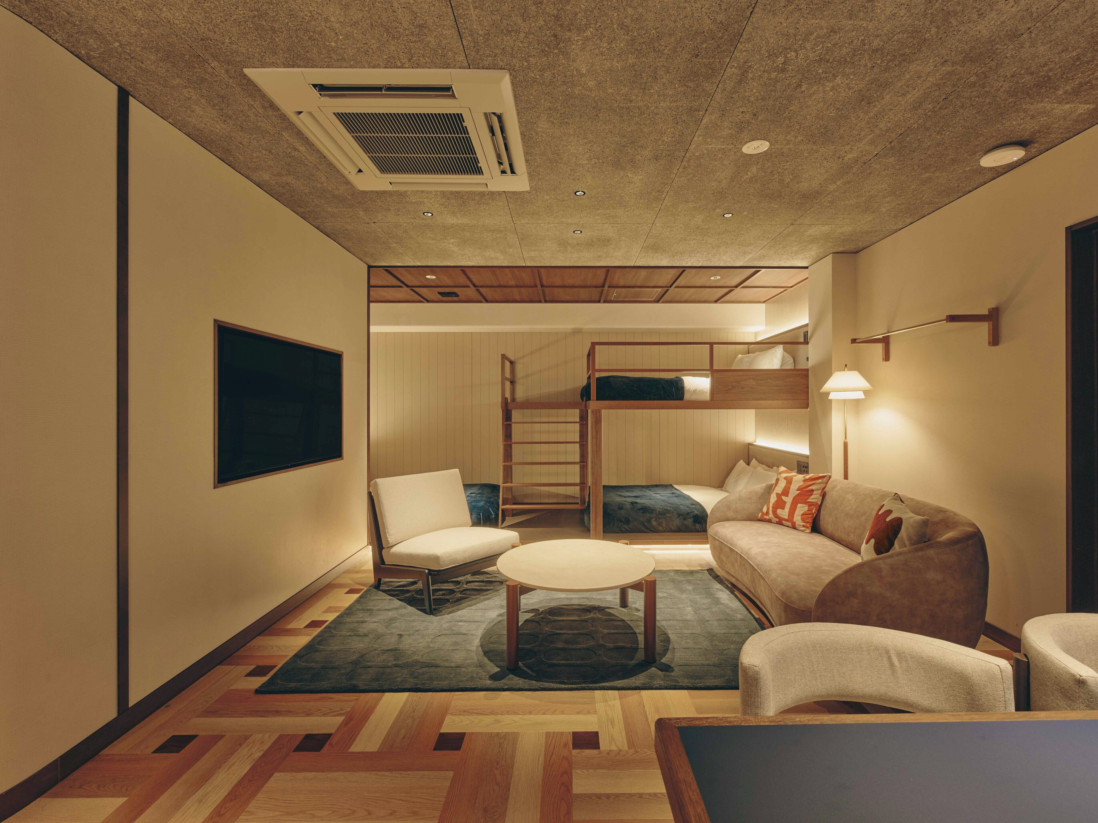 A compact hotel room with a bunk bed, a sofa, a chair, a small table, and a ceiling vent.