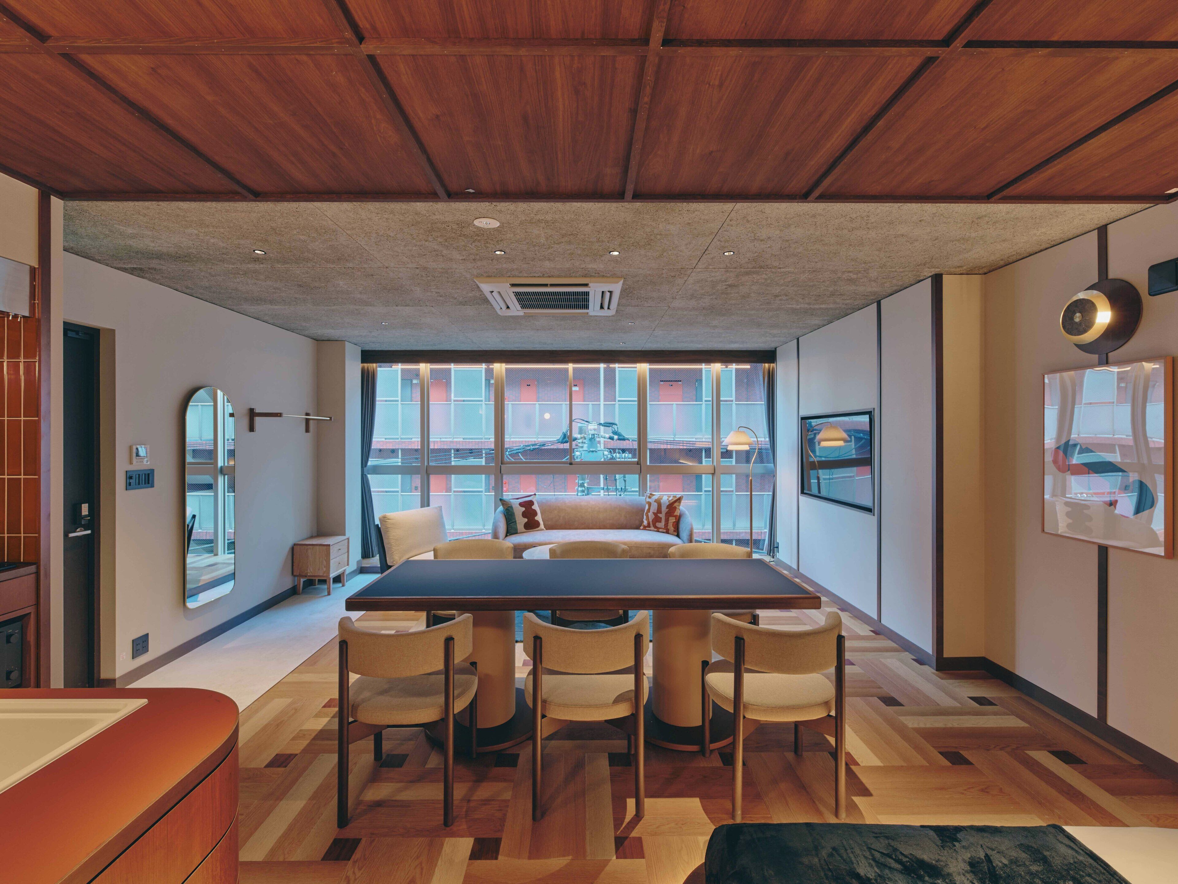 A modern hotel room with a large dining table, four chairs, a sofa, and a view of the cityscape through large windows.