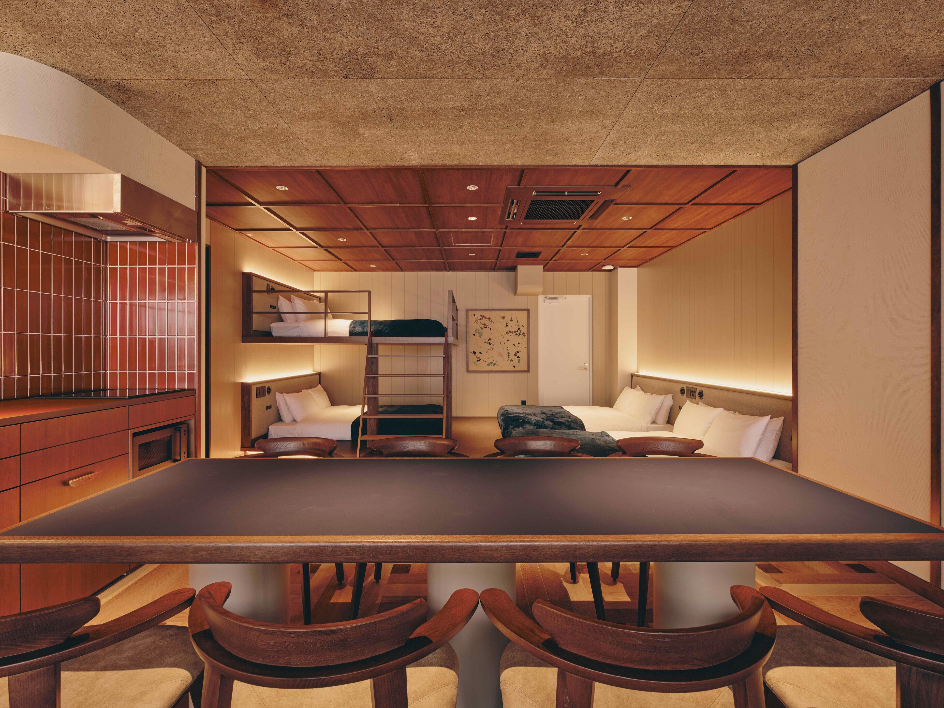 A modern hotel room with a large wooden table, four wooden chairs, a bunk bed, and a small kitchenette.