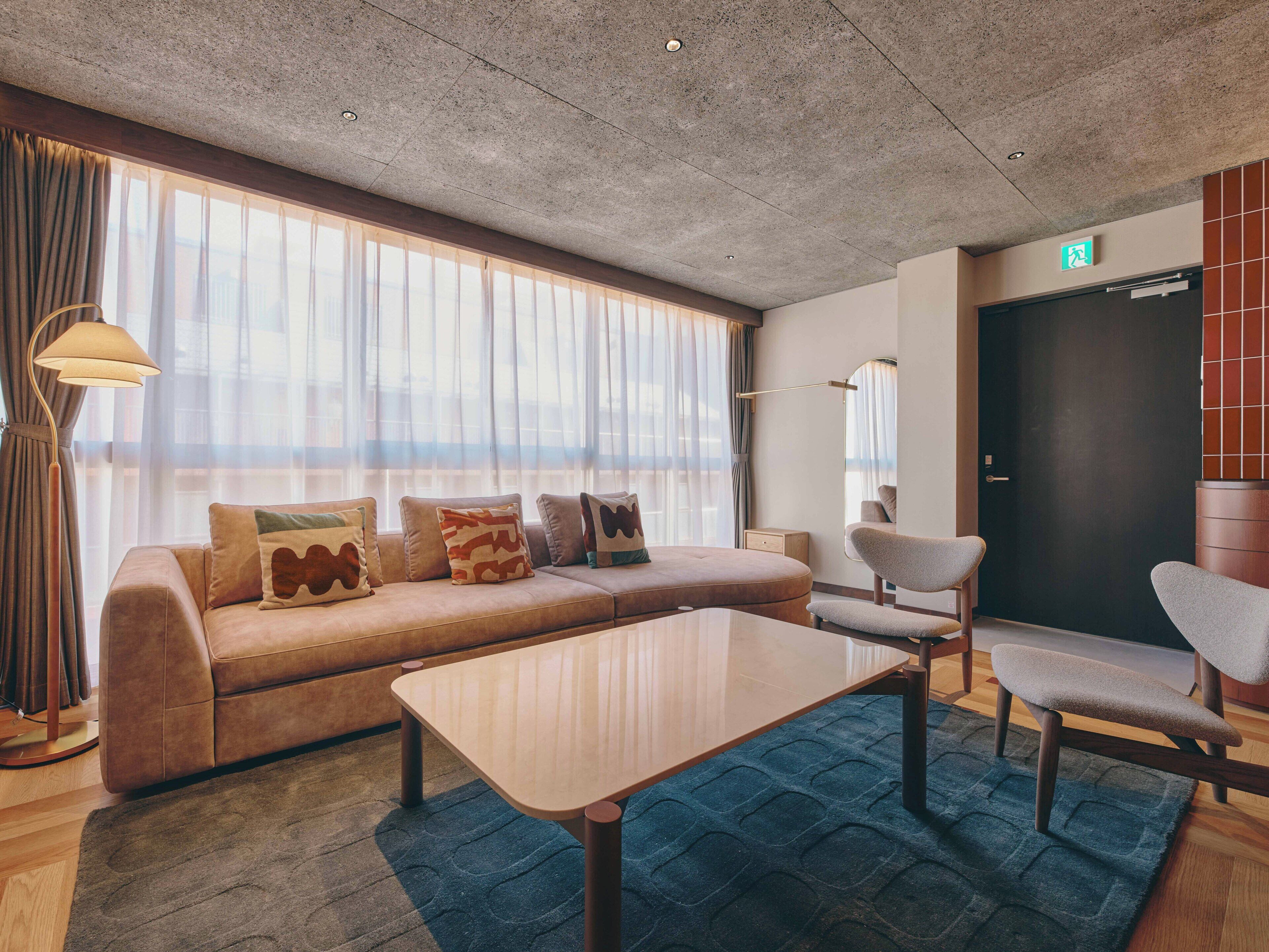 A modern hotel room with a sofa, armchairs, a coffee table, and a large window with sheer curtains.