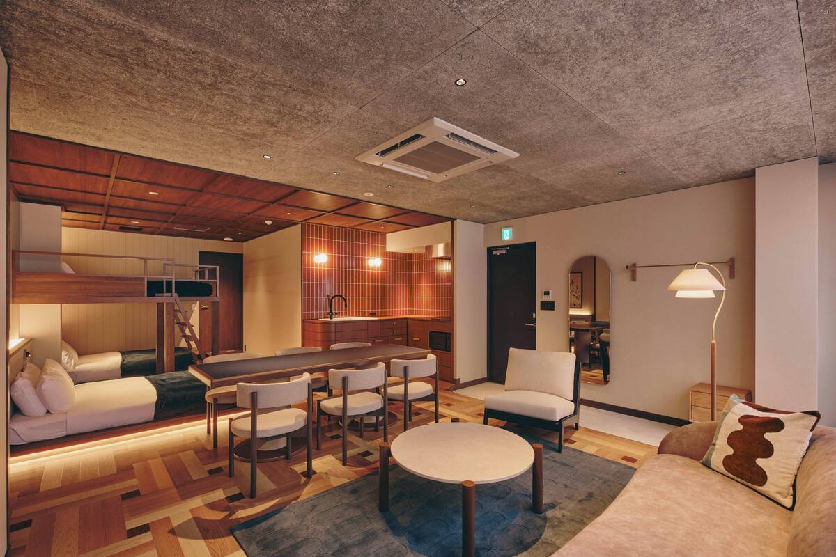 A modern hotel room with a bunk bed, a dining area with a long table and chairs, a sofa, a round coffee table, and a small kitchen area.