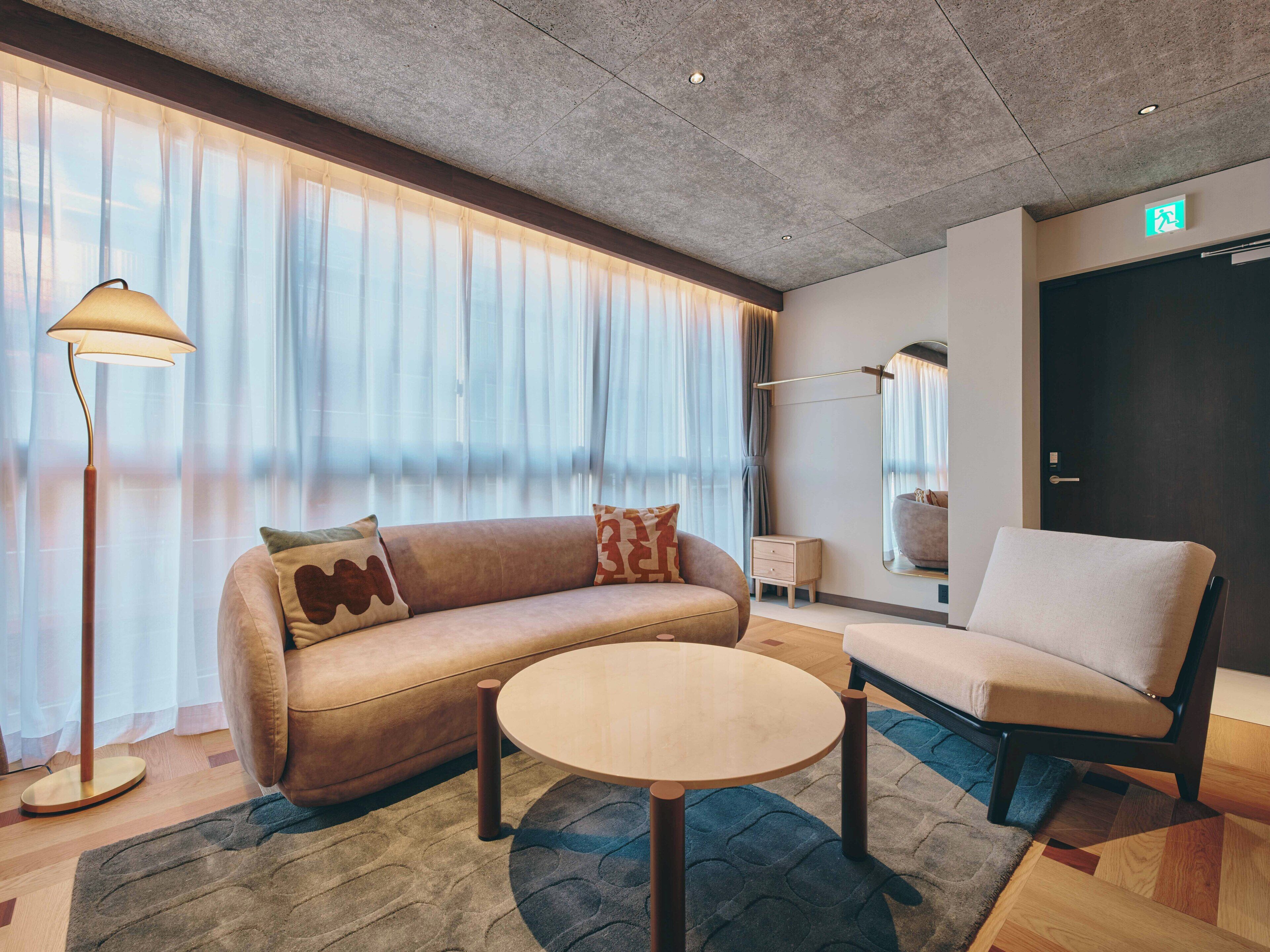 Una habitación de hotel moderna con sofá, sillón, mesa redonda, lámpara de pie y ventanas amplias con cortinas traslúcidas.