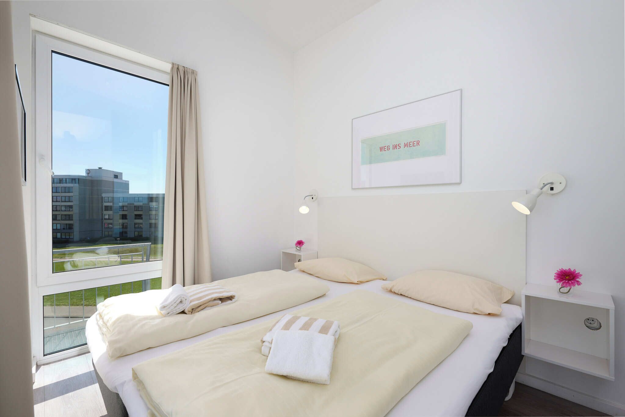 A hotel room with two beds, a large window with a view of buildings, a framed sign on the wall, and bedside lamps.