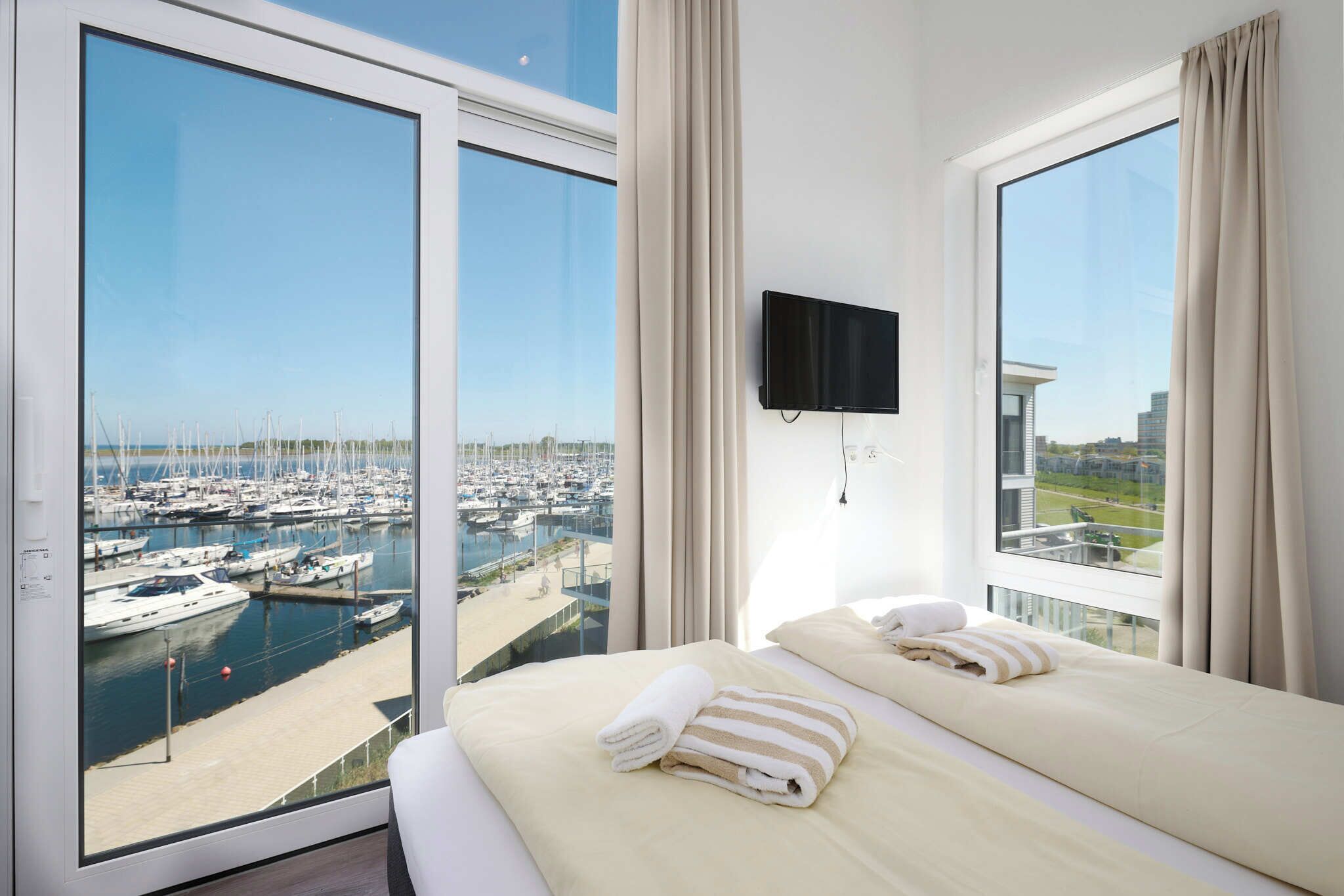 Une chambre d’hôtel dotée d’une grande fenêtre donnant sur un port de plaisance, d’un lit avec des serviettes et d’un téléviseur fixé au mur.