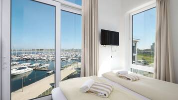 Une chambre d’hôtel dotée d’une grande fenêtre donnant sur un port de plaisance, d’un lit avec des serviettes et d’un téléviseur fixé au mur.