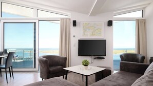 A modern living room with a large sofa, two armchairs, a coffee table, and a flat-screen TV mounted on the wall. There is a large window with a view of the beach and a map on the wall. - Beachfront Penthouse Apartment in Marina Wendtorf with Stunning Sea View (Wendtorf)