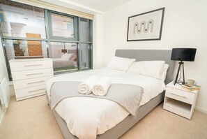 A bedroom with a bed, a bedside table, a lamp, a window, and a wall-mounted picture. - Bright 2 Bed Apartment in Nottingham Centre (Nottingham)