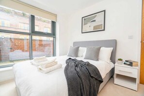 Ein ordentlich bezogenes Bett mit weißen Leinentüchern und einer grauen Decke, ein Nachttisch mit Büchern und einer Pflanze sowie ein Fenster mit Blick auf ein Backsteinhaus.