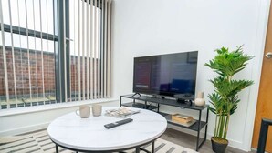 A modern living room with a TV, a round table, a coffee table, a potted plant, and a window with blinds. - Charming 2BED Apartment in the Heart of Nottingham (Nottingham)