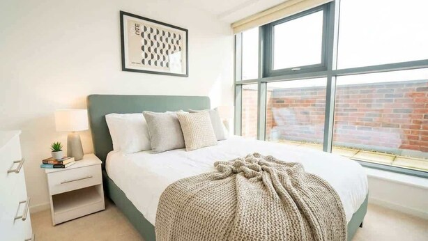 A bedroom with a bed, a nightstand, a lamp, a framed picture, and a window with a view of a brick building. - Charming 2BED Apartment in the Heart of Nottingham (Nottingham)