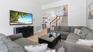 Una sala de estar con un televisor de pantalla plana grande, sofás grises, una mesa de centro y una escalera que lleva a un nivel superior.