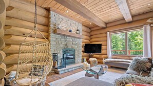 L’intérieur d’une cabane en rondins, avec une cheminée en pierre, un fauteuil suspendu, un téléviseur et une grande fenêtre.