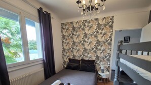 Une chambre avec un grand lit, une fenêtre avec des rideaux, un radiateur, un lustre, un lit superposé et un mur orné d’un motif de feuilles tropicales.