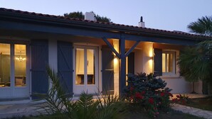 Une maison dotée d’un porche abrité, de grandes fenêtres et d’un jardin agrémenté de fleurs rouges.