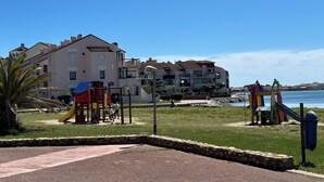 Ein Spielplatz mit Rutschen und Klettergerüsten, eine Mauer aus Stein und ein Gebäude mit Balkonen mit Blick auf ein Gewässer.