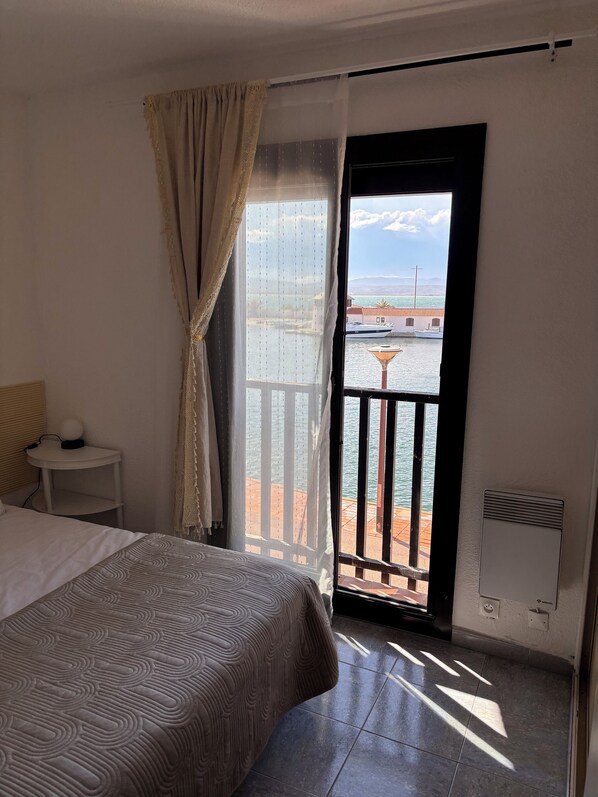A hotel room with a bed, a small bedside table, a window with a view of the sea, and a balcony with a railing.