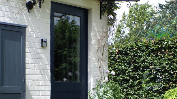 Uma casa com porta azul escura, um pequeno alpendre e um jardim com diversas plantas.