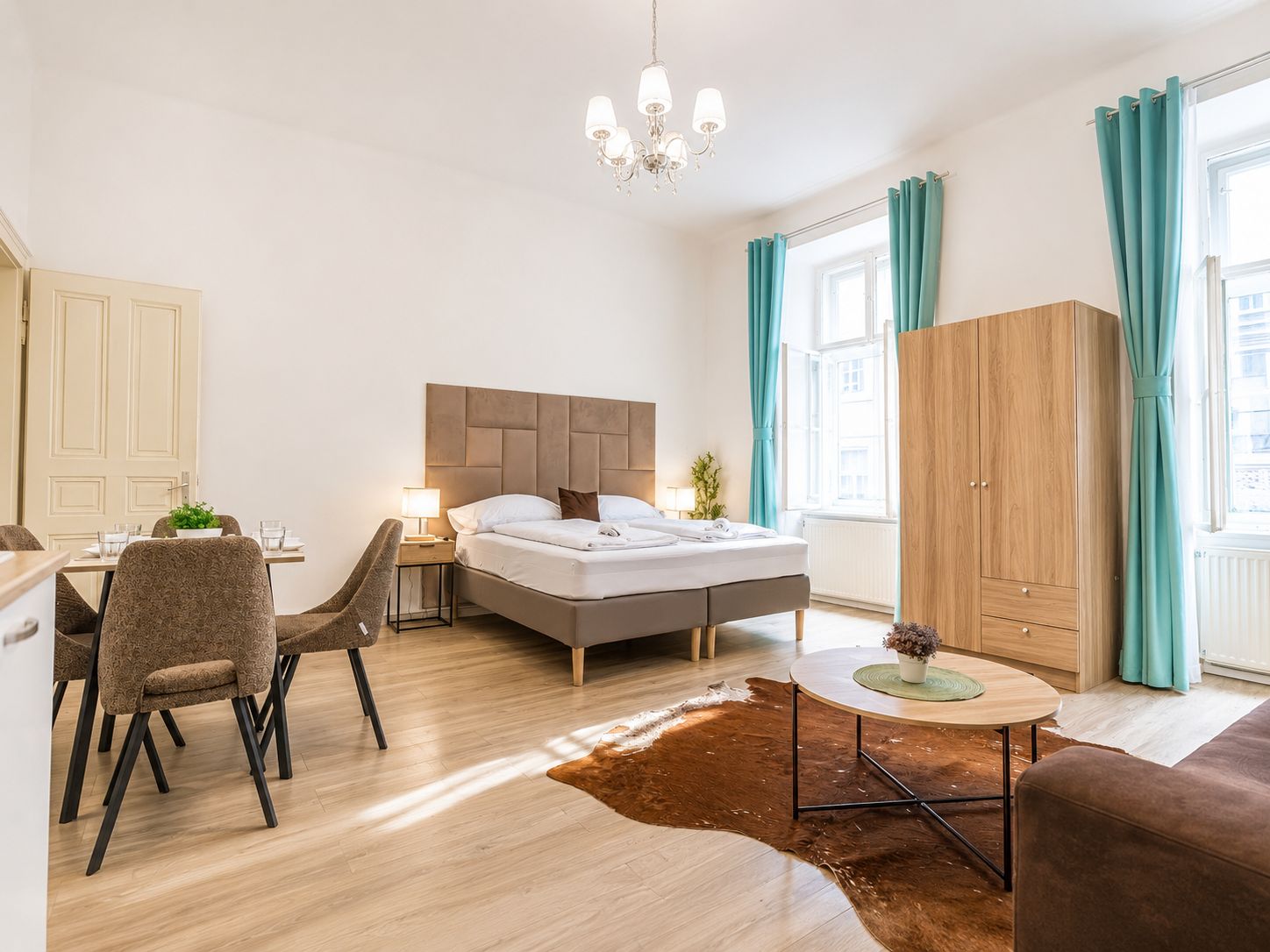 Hotellihuone, jossa on sänky, työpöytä tuoleineen, pieni pöytä, vaatekaappi ja ikkuna, jossa on verhot.
